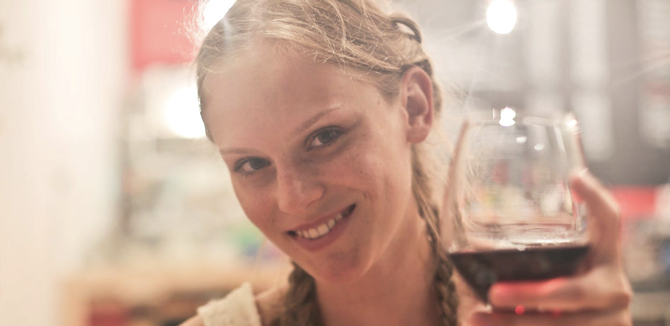 Young woman smiling into the camera while holding up a glass of wine (image representing a sugar baby on a date with a sugar daddy to tie into topic of sugaring and sugar dating)