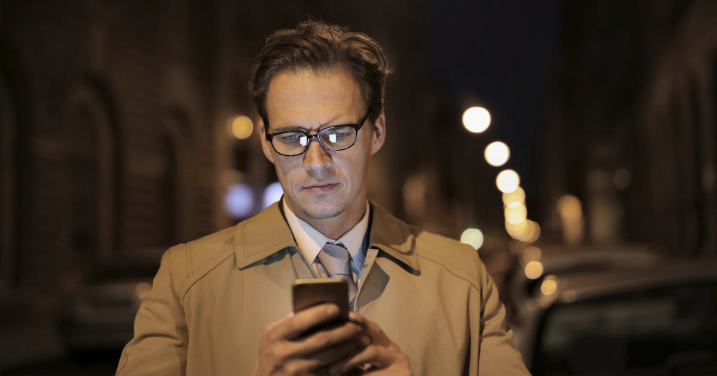 A man checks his phone on a dark street (Representing the return of The Erotic Review